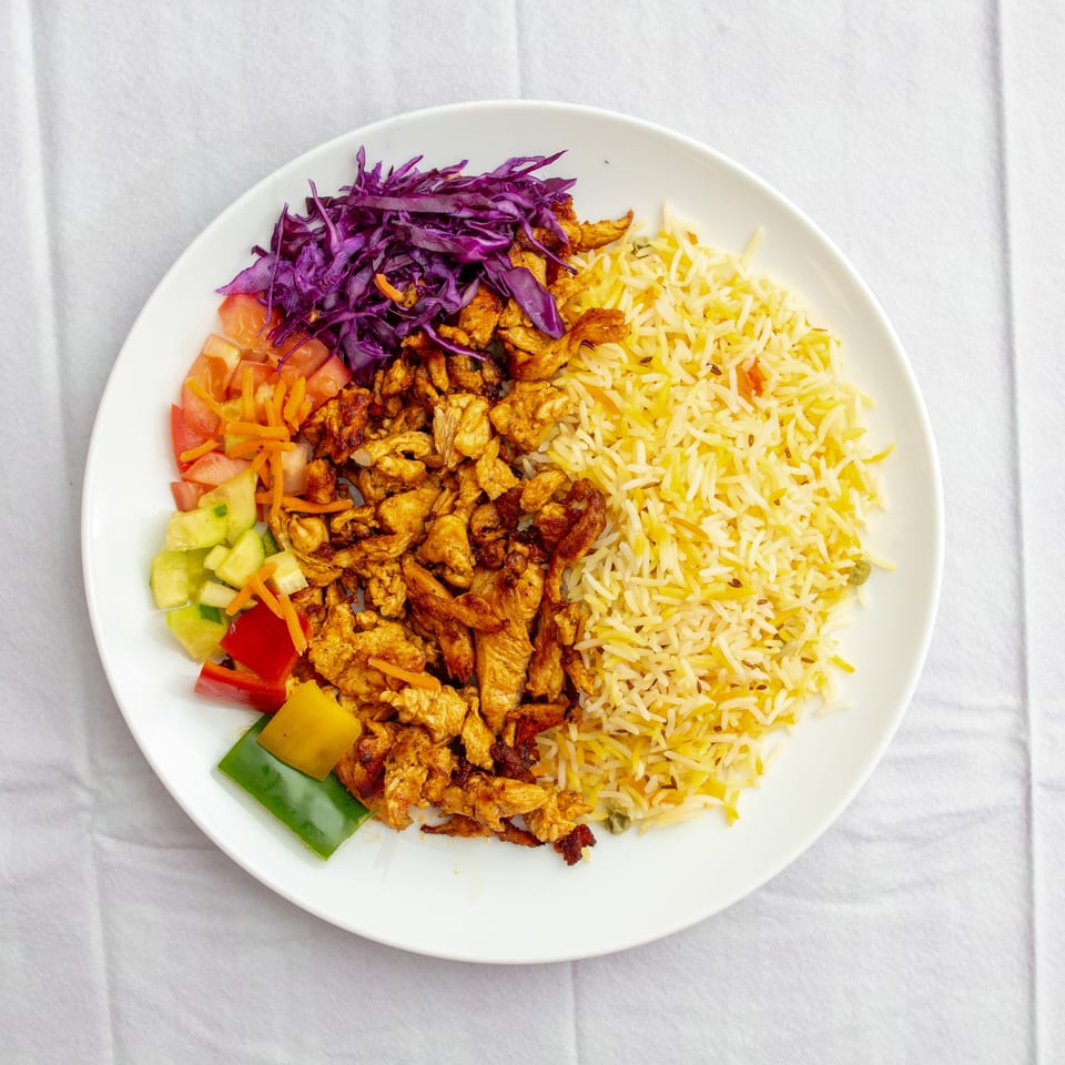 Plate of seasoned chicken, yellow basmati rice, shredded purple cabbage, and chopped vegetables on a white tablecloth.