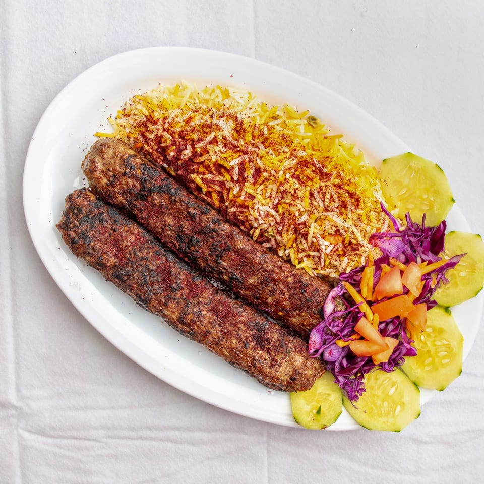 Grilled kebabs served with spiced yellow rice, sliced cucumbers, tomatoes, carrots, and purple cabbage on a white plate.
