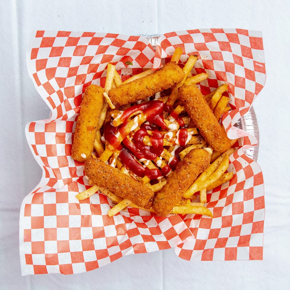 Crispy fried cheese sticks and seasoned fries topped with ketchup and creamy sauce in a red-and-white checkered tray.